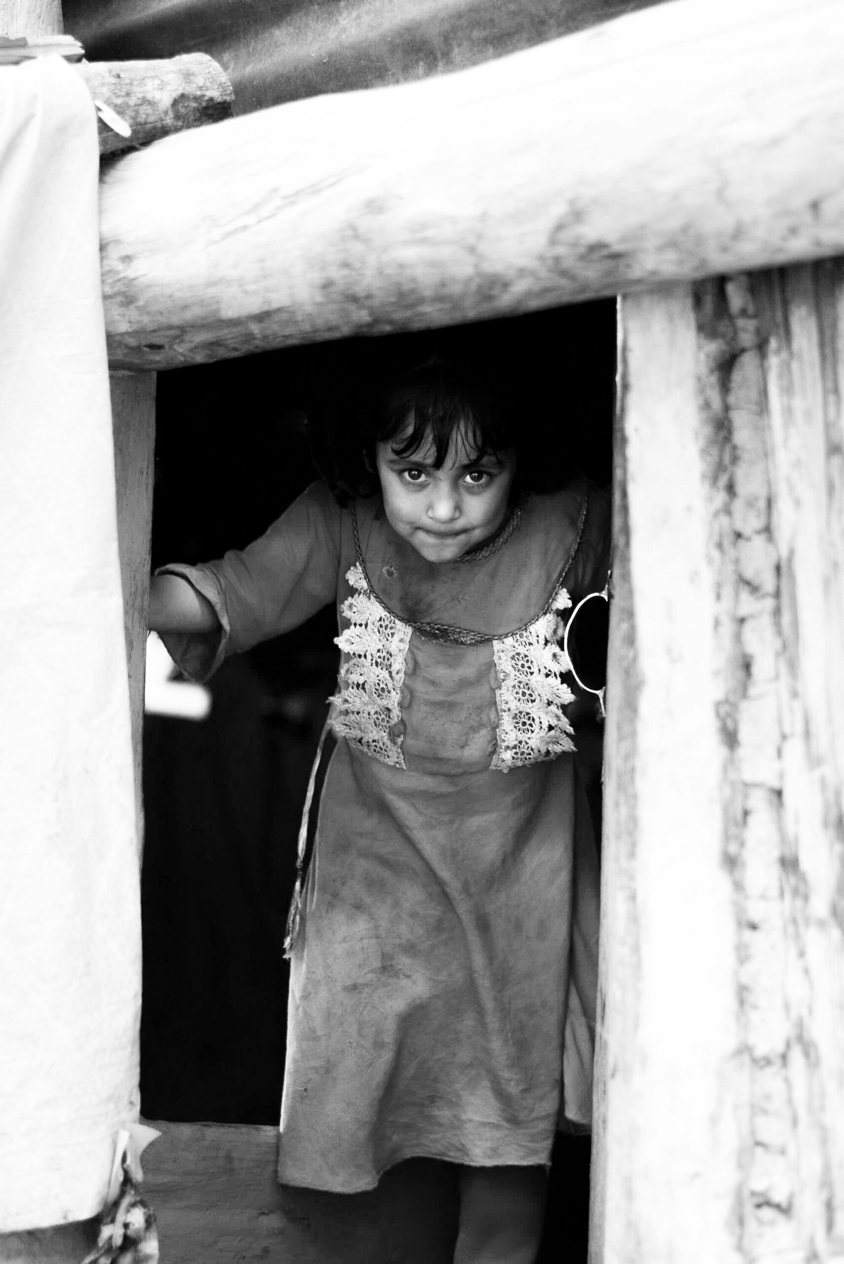 Monochrome portrait of a young child peering out from a rustic shelter in Dardpora.